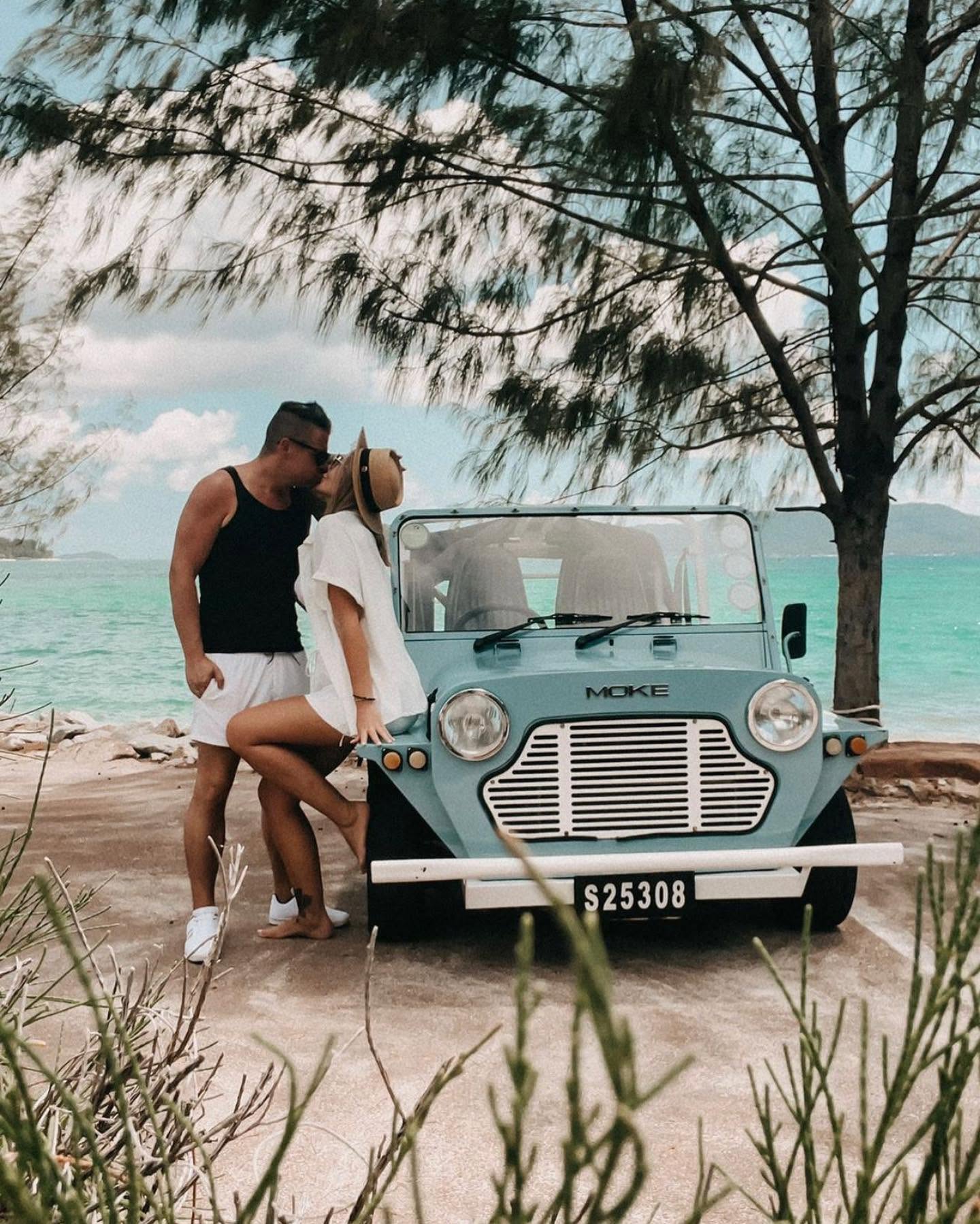 A couple standing beside a pale blue Mini Moke on a concrete slipway at the edge of a turquoise bay, casuarina branches overhead