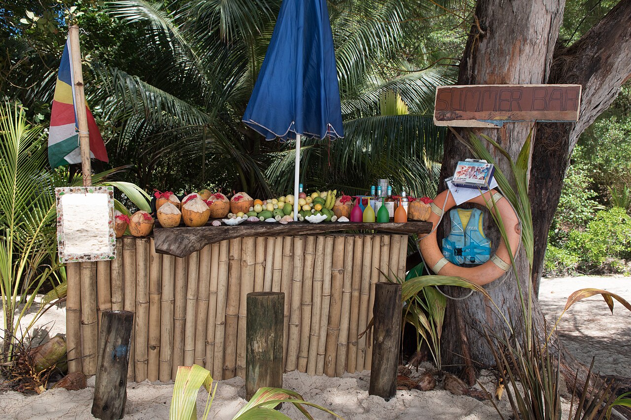 A beach bar counter at Anse Intendance on Mahé, coconuts and fruit set out along the bamboo and the Seychelles flag flying behind it.