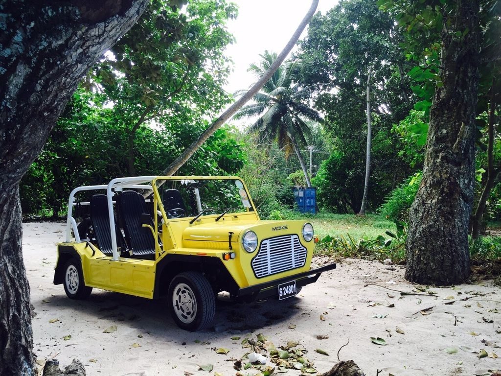 A yellow Mini Moke parked on white sand between takamaka trunks with coconut palms behind
