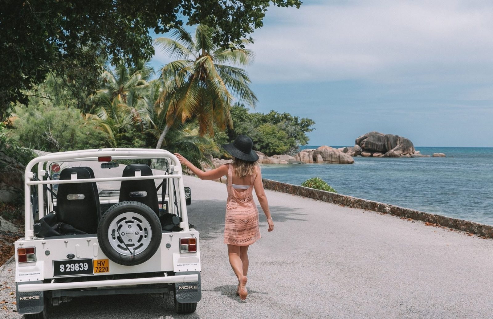 A white modern Mini Moke seen from behind with its spare wheel on the tailgate, on a coast road with granite boulders and the sea beyond