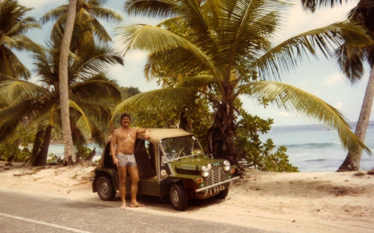 A 1980s photograph of a man in swimming shorts standing beside an olive green Mini Moke on a coast road under coconut palms