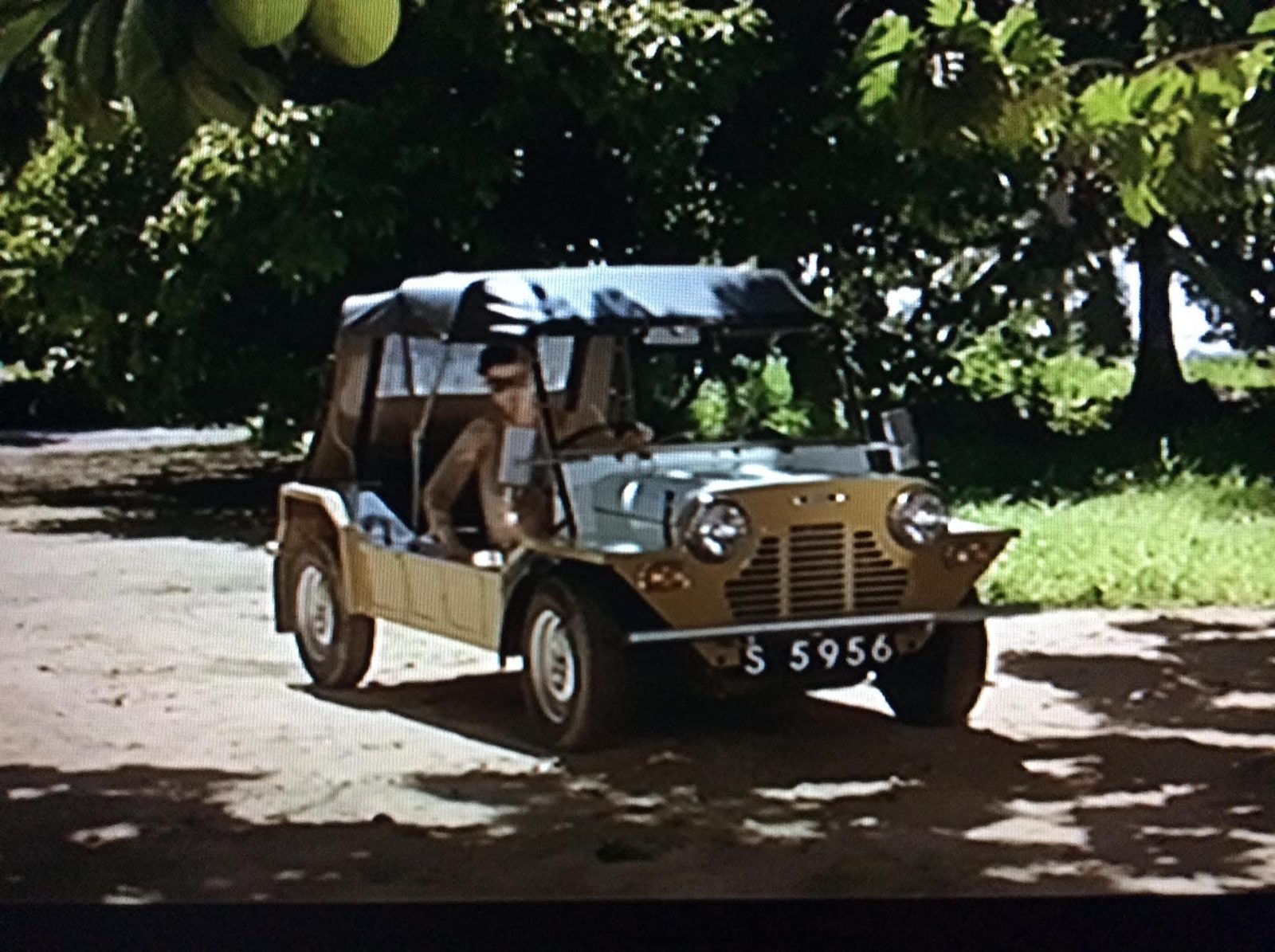 A gold classic Mini Moke with a black canvas top on a sandy road under takamaka trees