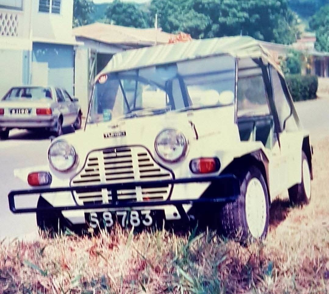 A cream Mini Moke with a canvas roof parked on a Mahé street in front of an older saloon and a shopfront