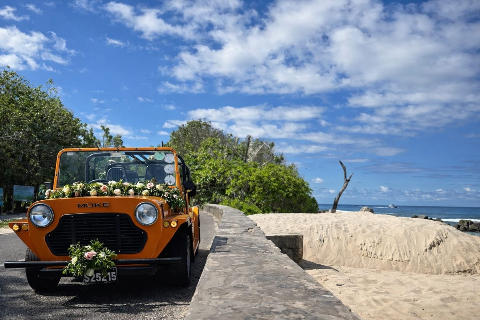 An orange Mini Moke dressed with white flowers on a low wall above a beach