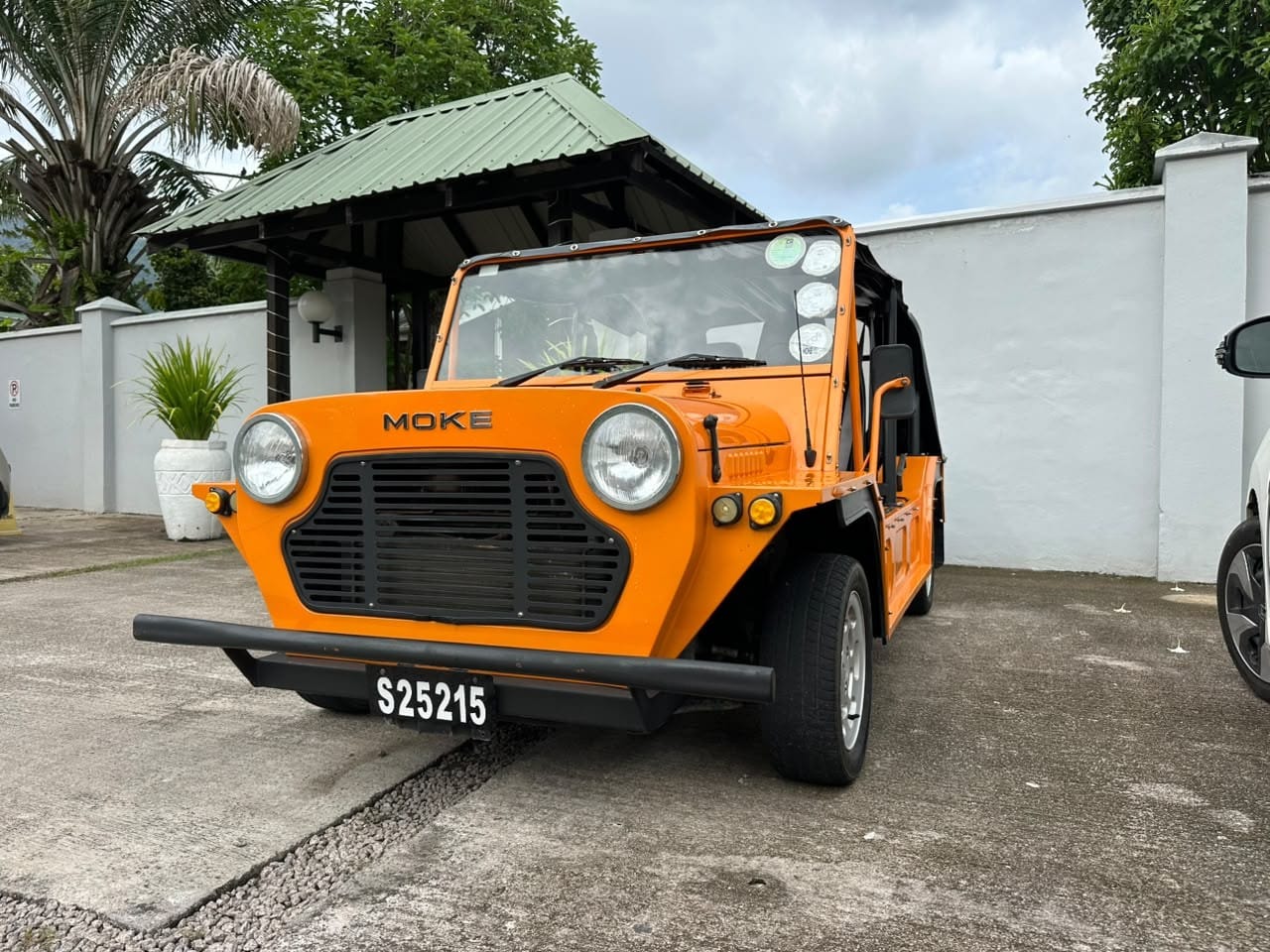 An orange Mini Moke with the MOKE badge on its grille, waiting at the gate of a white-walled villa