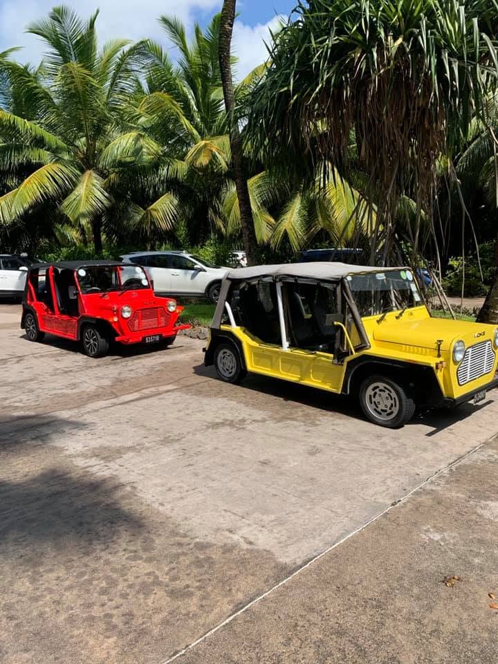 A red Mini Moke and a yellow one parked side by side on a concrete yard under palms