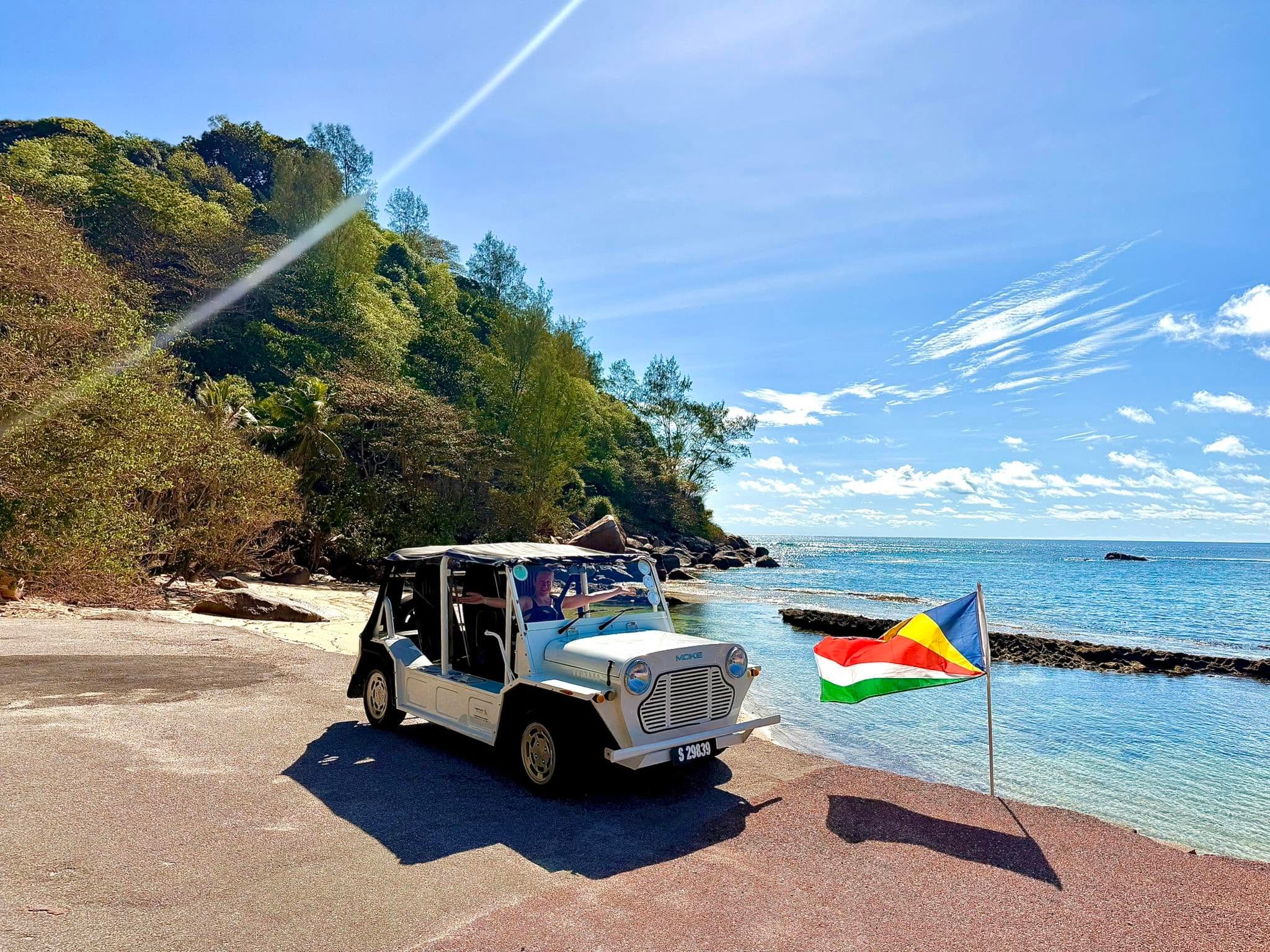 A cream Mini Moke parked on a slipway at the water's edge, a Seychelles flag beside it and a wooded headland across the bay