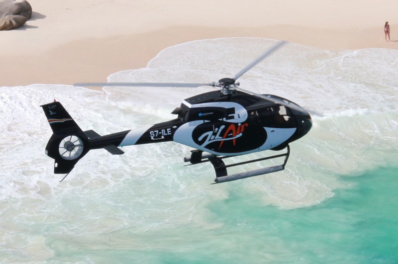 A Zil Air helicopter flying low over a turquoise shallow bay and an empty white beach