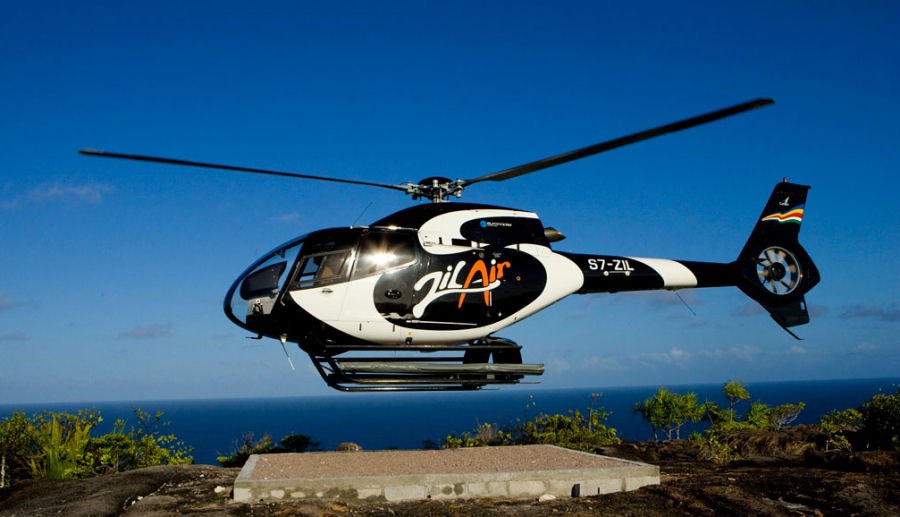 A Zil Air helicopter airborne beside a coastal helipad with open sea behind