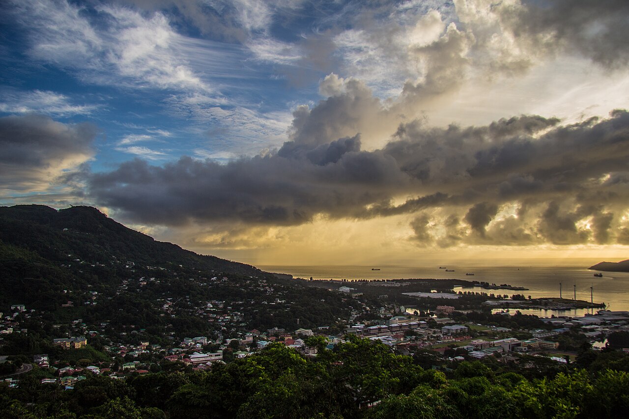 Victoria and its harbour at sunrise, seen from the hills of Mahé, with ships lying off the town and the open ocean beyond.