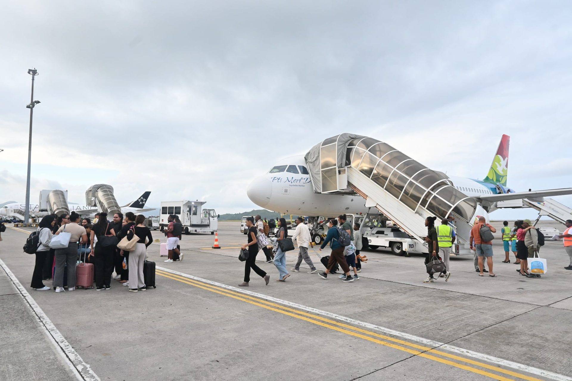 Passengers walking in across the apron with their bags, an Air Seychelles aircraft on the stand behind with the airstairs down