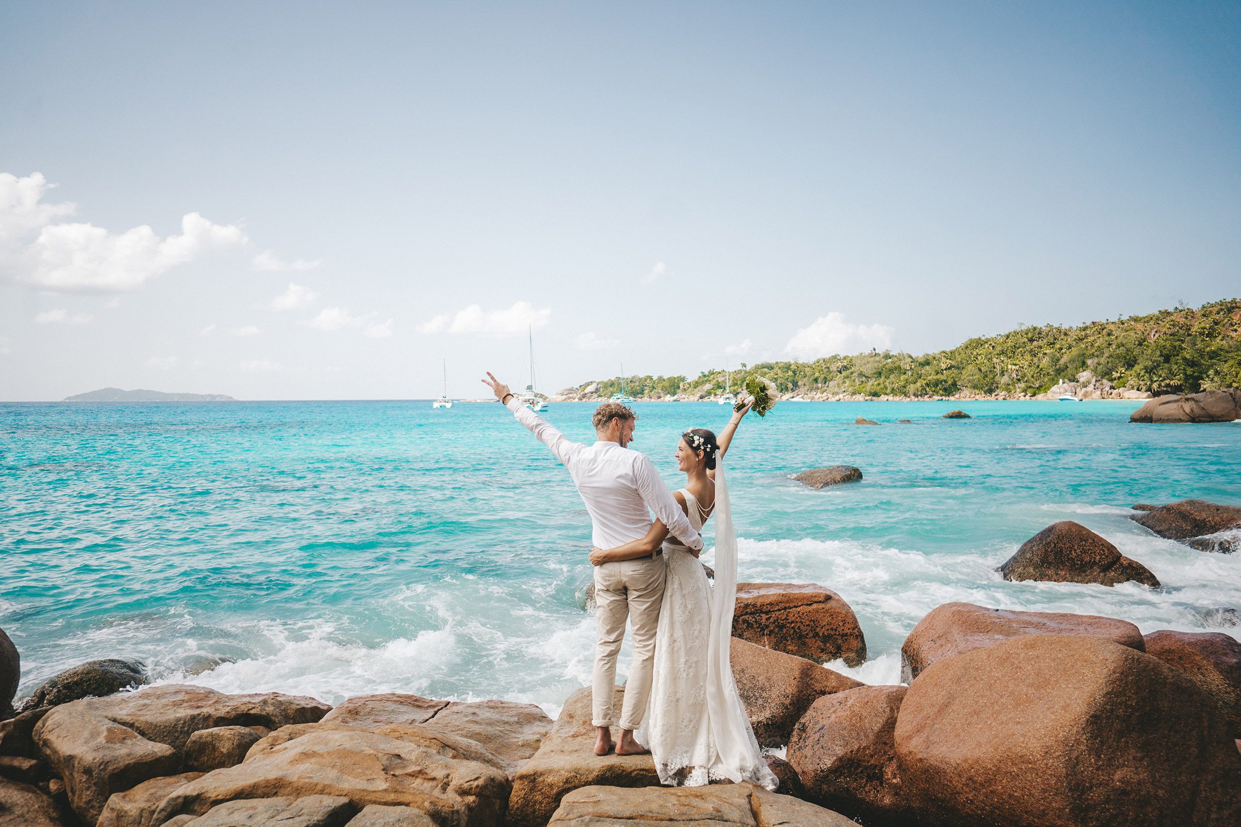 The couple on the rocks with their arms up, a yacht on the turquoise water behind