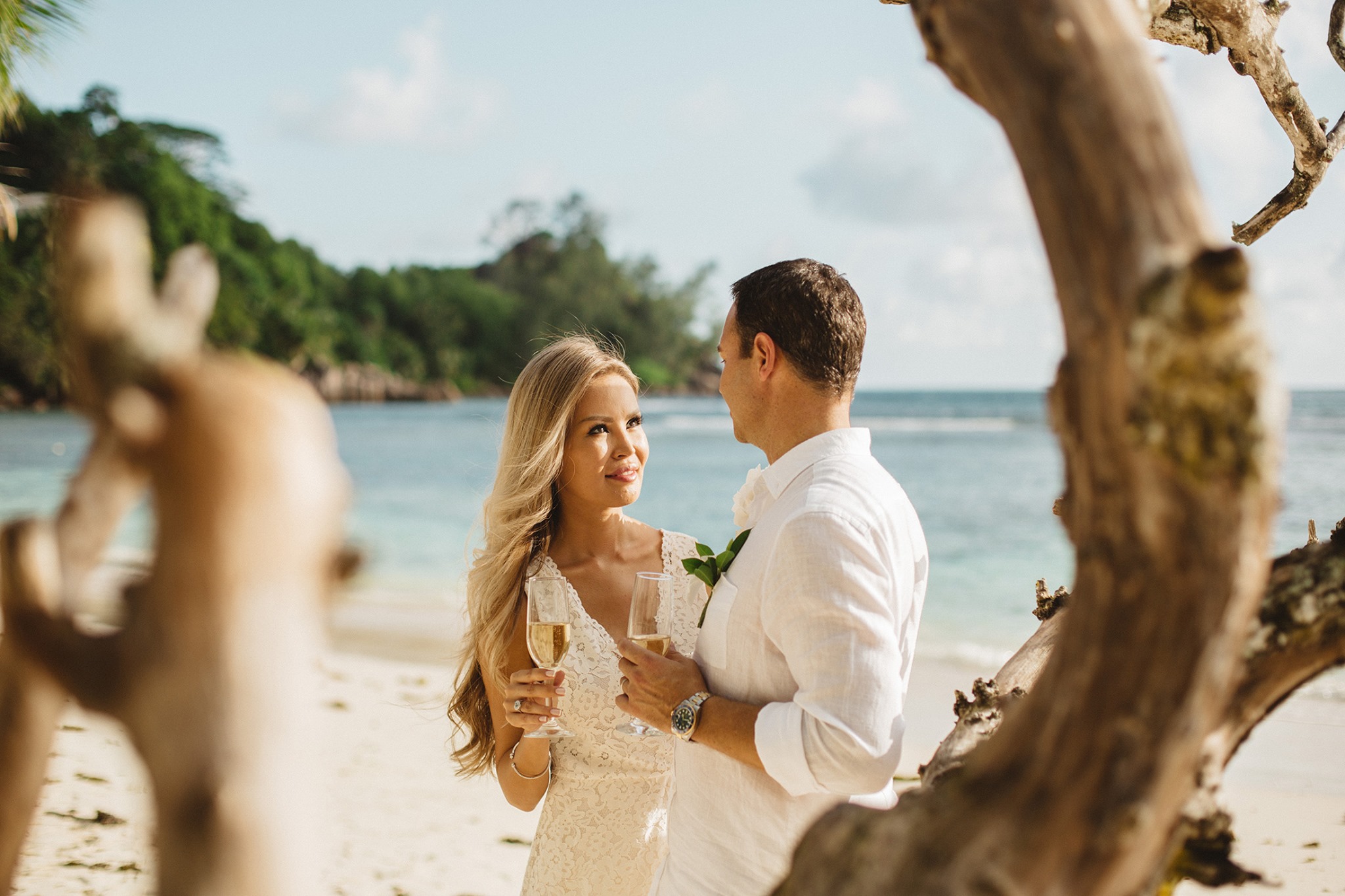A bride in a lace gown and a groom in white linen, framed by a bleached driftwood branch, the lagoon behind them