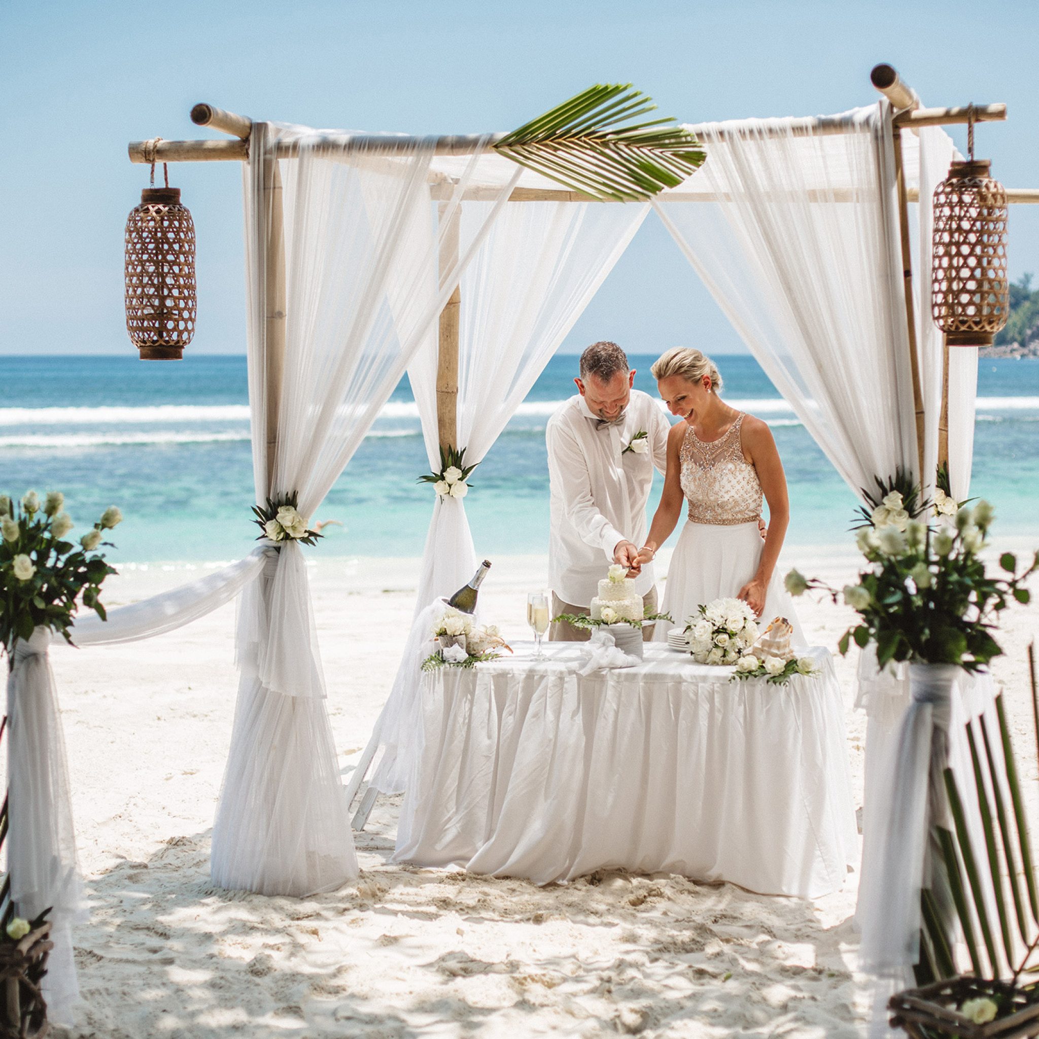 An empty ceremony set on the sand: a bamboo frame, white drapes, lanterns and a dressed table