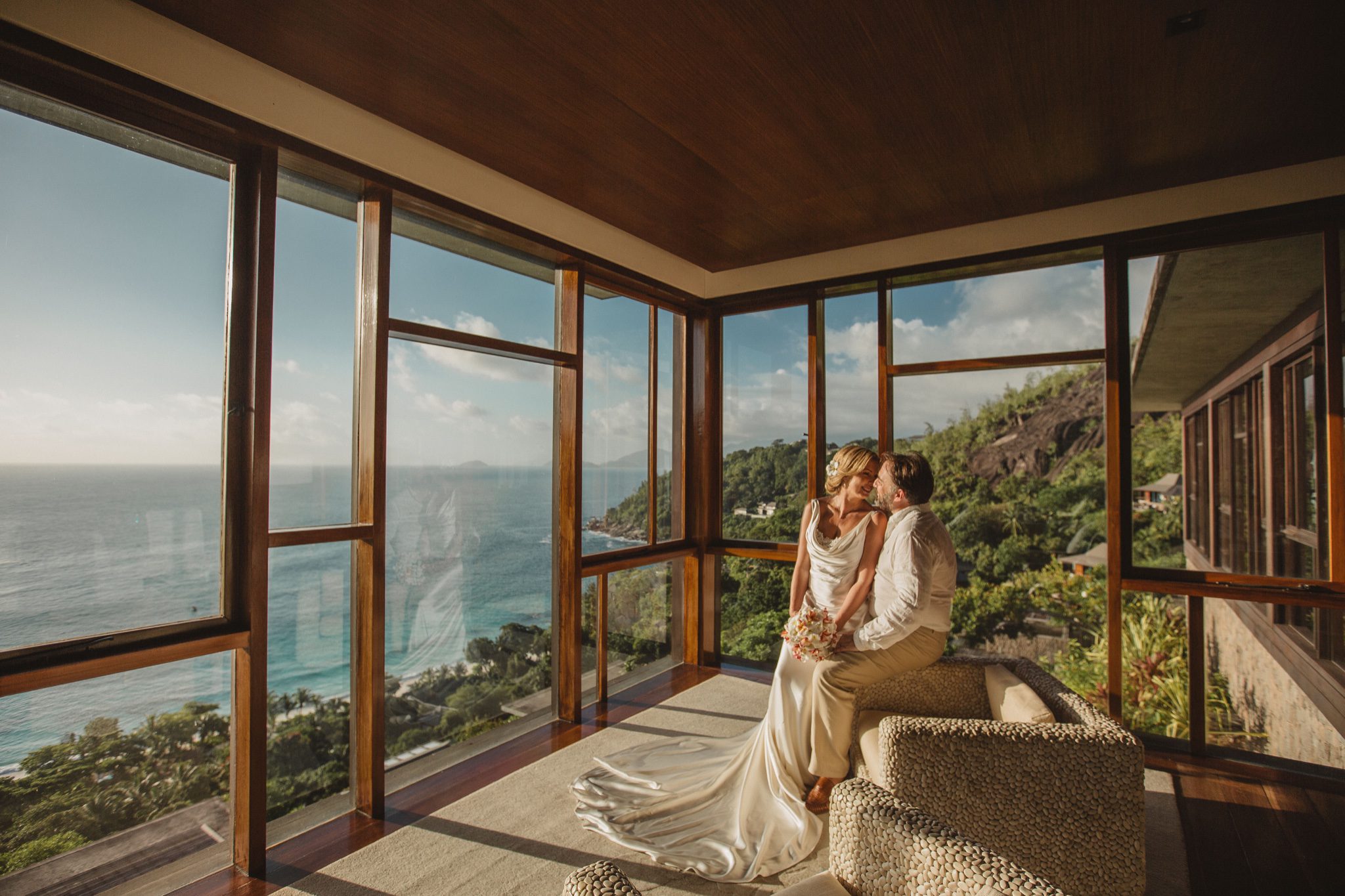 The couple sitting together in a glass-walled room looking over forest to the sea