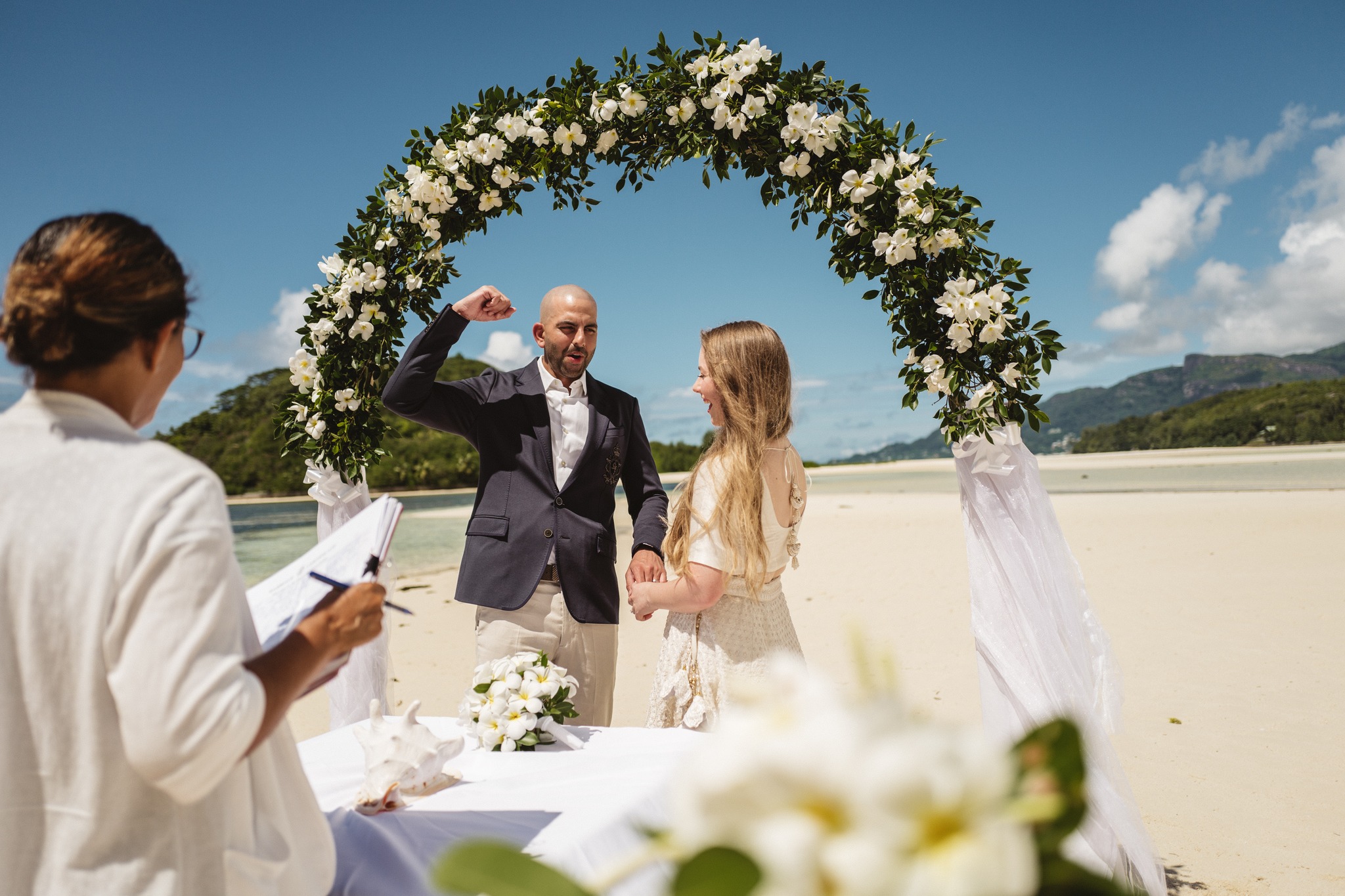 The registrar writing at a white table while the couple stand together under an arch of white flowers