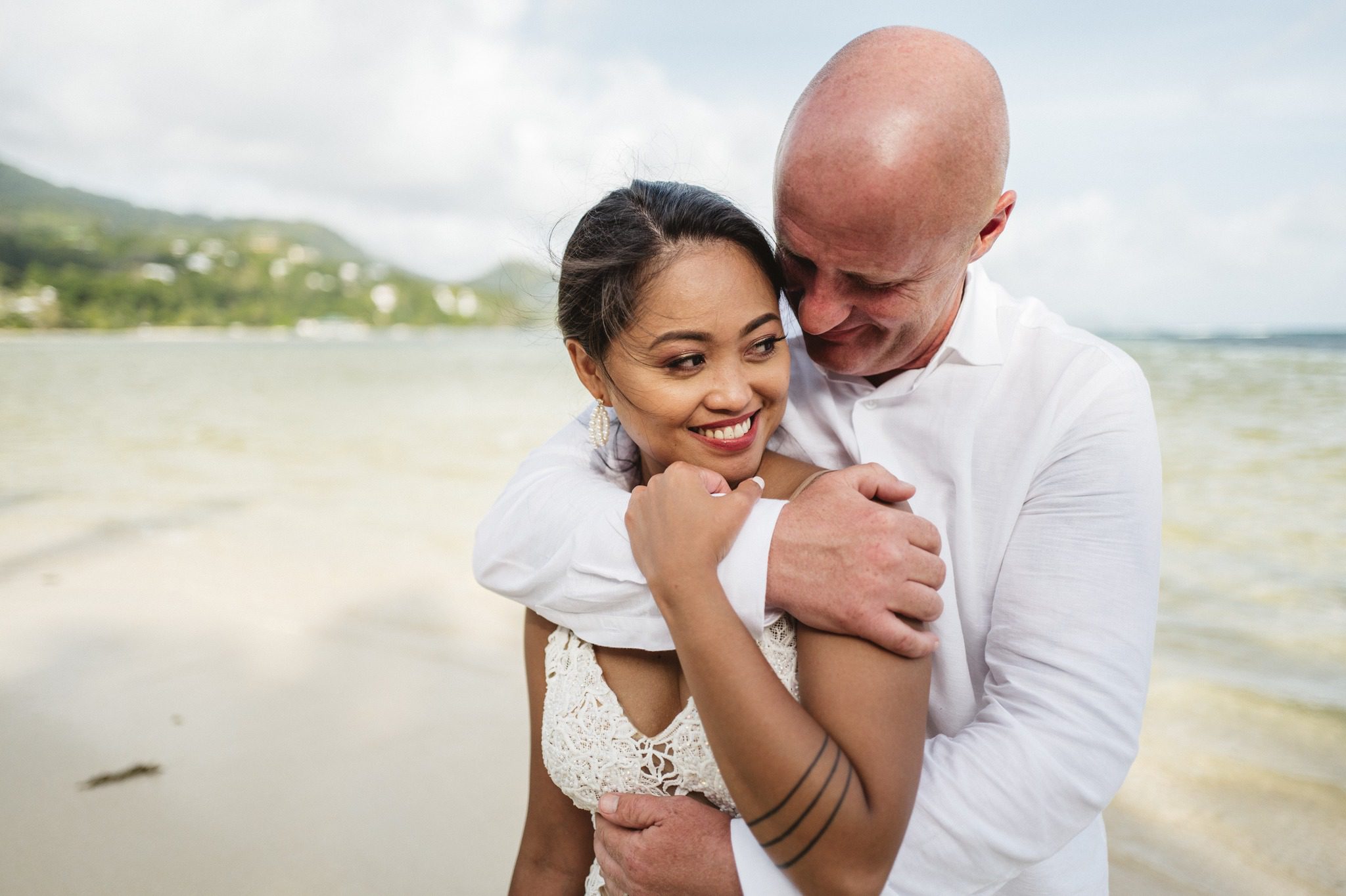 The bride tucked under the groom's arm, both smiling, the lagoon behind