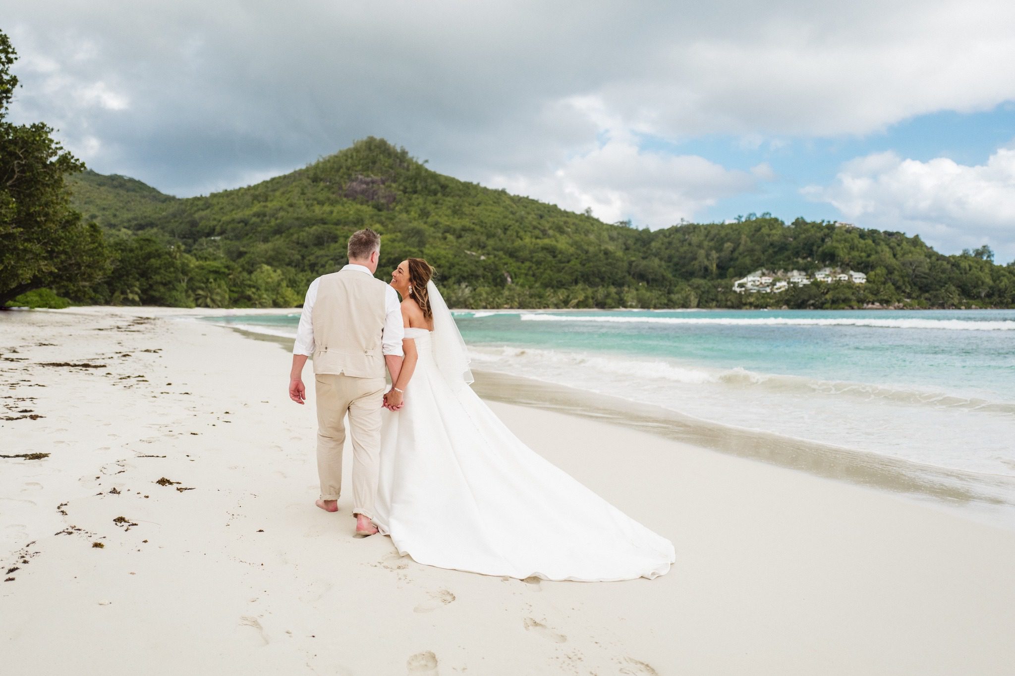 Bride and groom walking away down a wide empty beach, a green hill across the bay